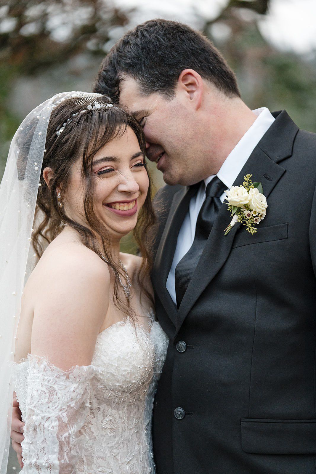 A joyful bride in a lace gown and veil laughs as her groom in a black tuxedo nuzzles her cheek outdoors.
