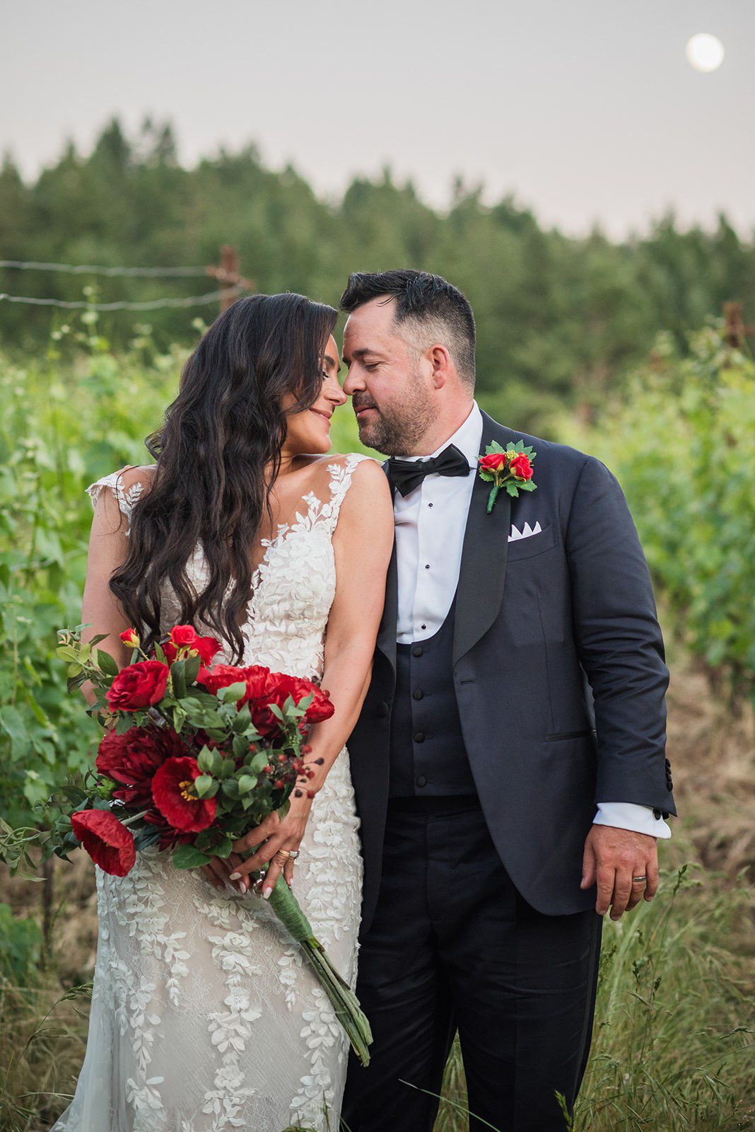 Bride and groom share a romantic moment in a vineyard, bride holding red bouquet, groom in black tuxedo.