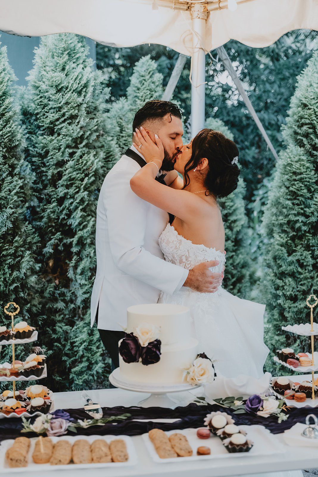 Bride and groom share a kiss behind their white wedding cake at an outdoor reception surrounded by evergreen trees.
