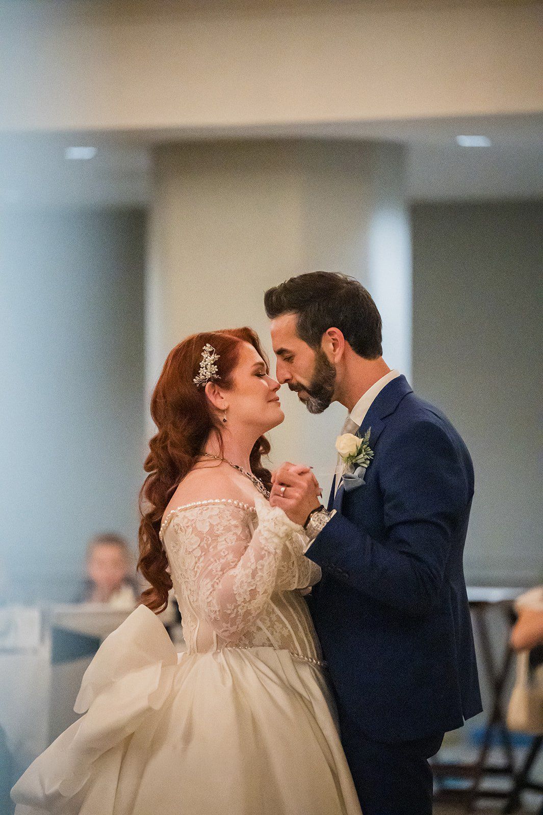Bride and groom share first dance, bride in lace off-shoulder gown, groom in navy suit with boutonniere.