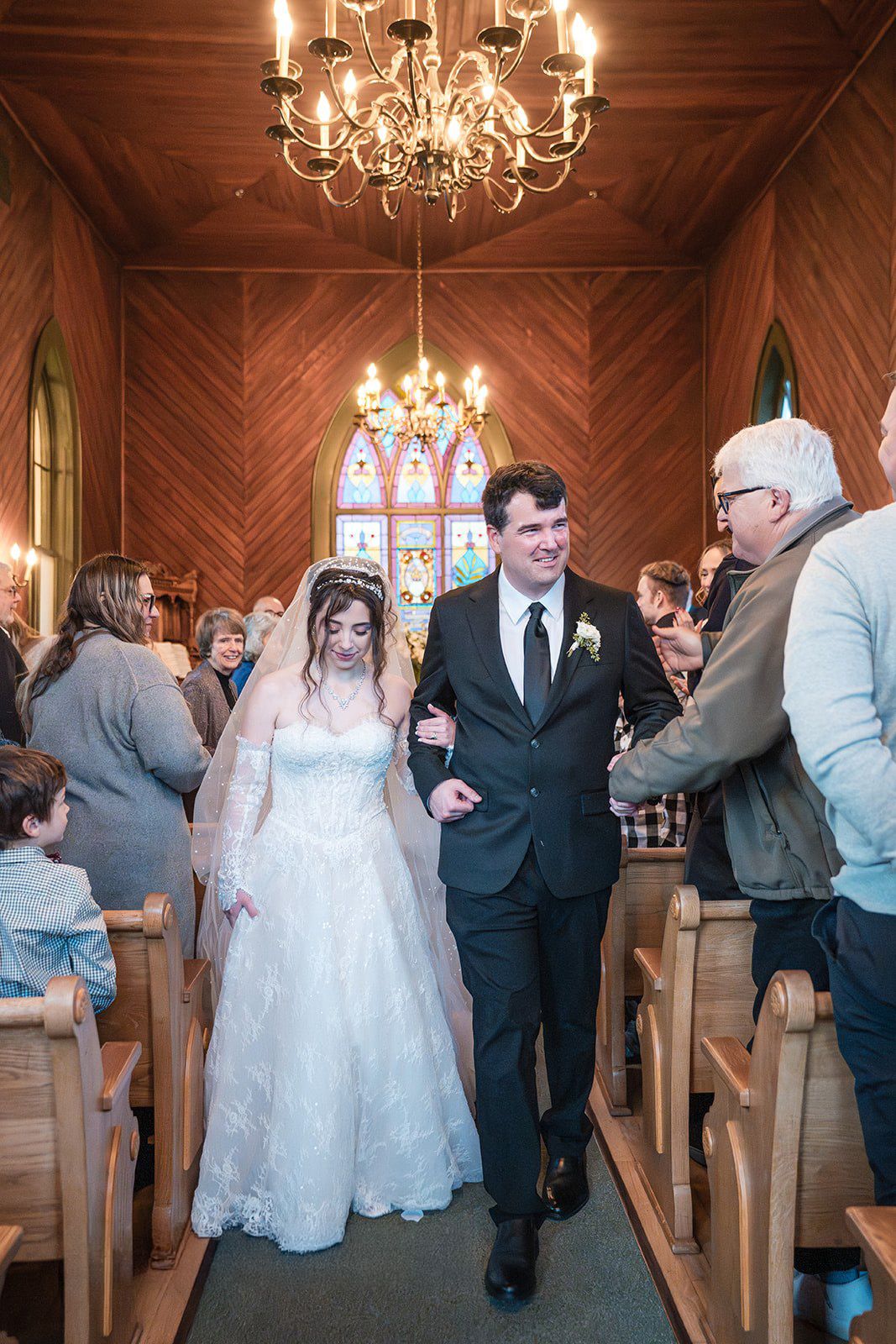 Bride and groom walk down chapel aisle after ceremony in elegant wood-paneled church with chandelier and stained glass.
