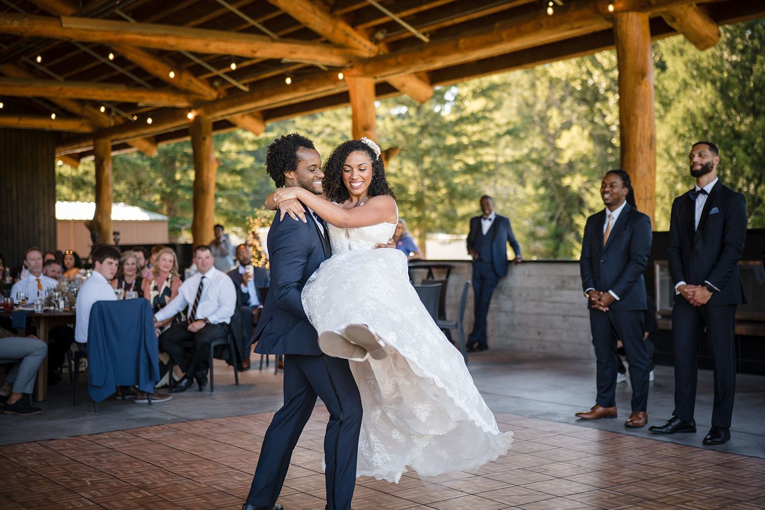 Groom lifts bride during first dance at outdoor wedding reception under string lights with guests watching.