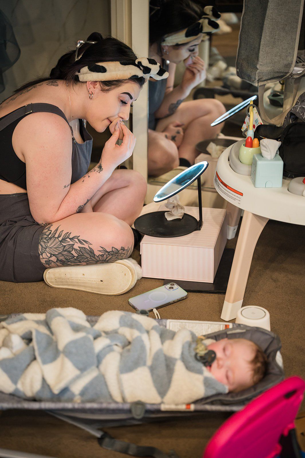 Tattooed mother applies makeup on floor beside sleeping newborn in bassinet, surrounded by vanity mirrors.