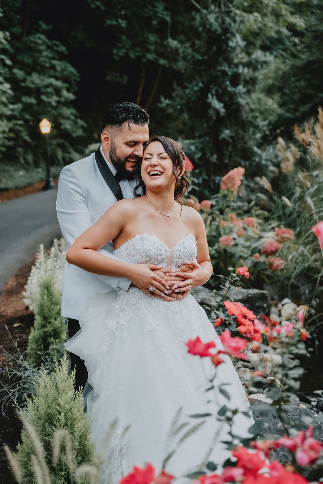 A joyful bride in a strapless gown laughs as her groom in a white tuxedo embraces her among colorful garden flowers.