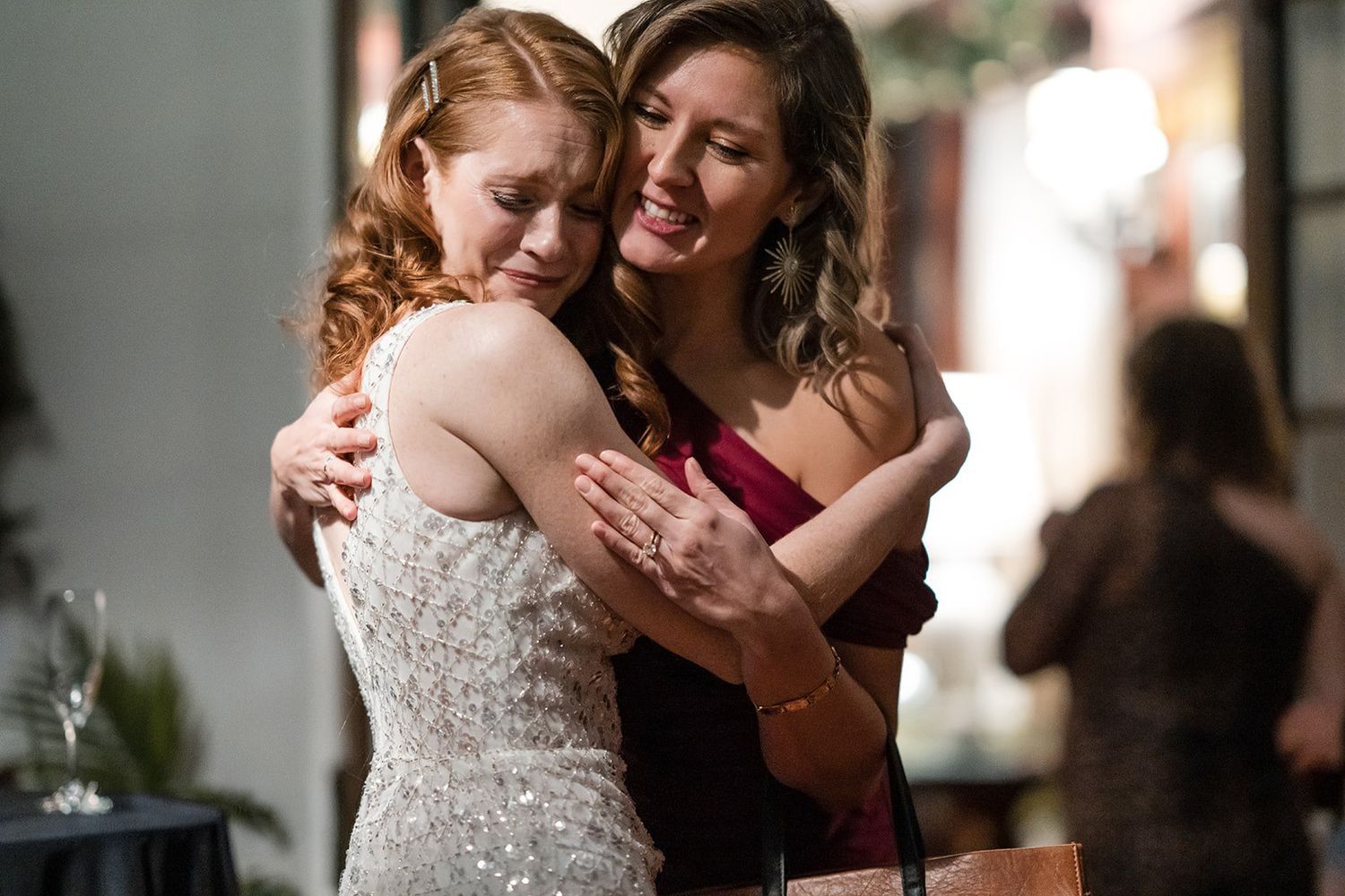 Two women share a warm embrace at a wedding, one in a white gown and the other in a red dress.