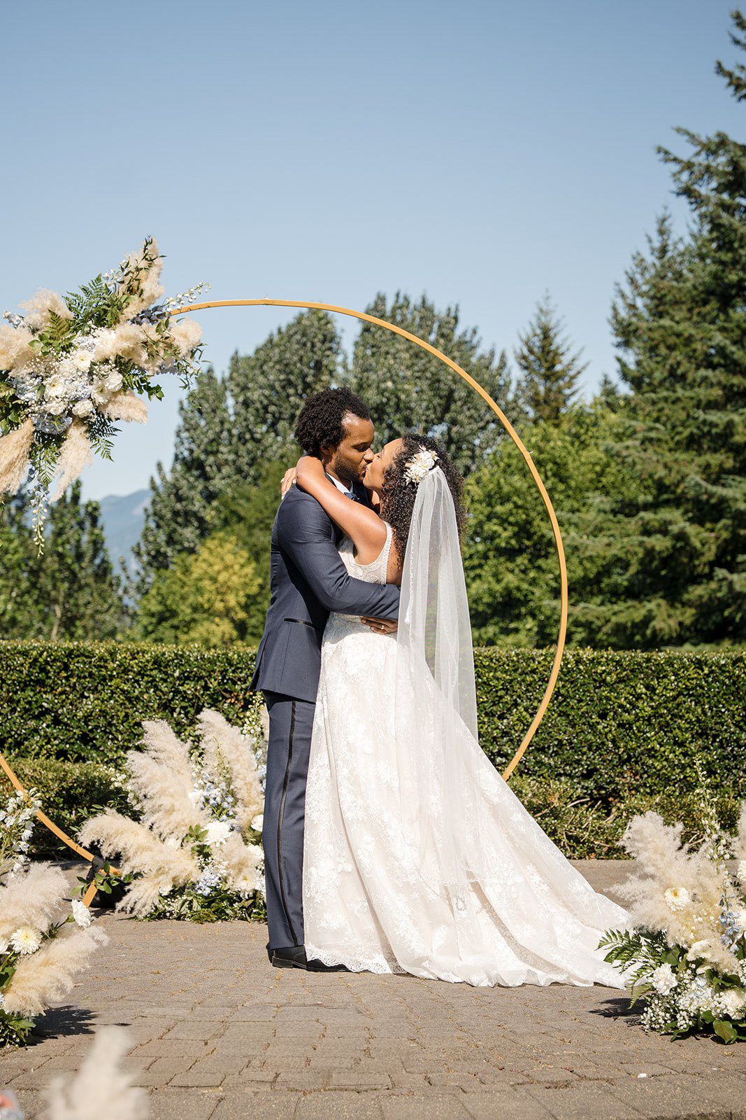 Bride and groom share first kiss under golden circular arch with pampas grass florals outdoors on sunny wedding day.