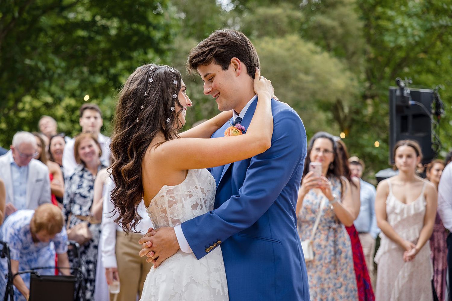 Bride and groom share their first dance outdoors, surrounded by smiling wedding guests in a lush garden setting.