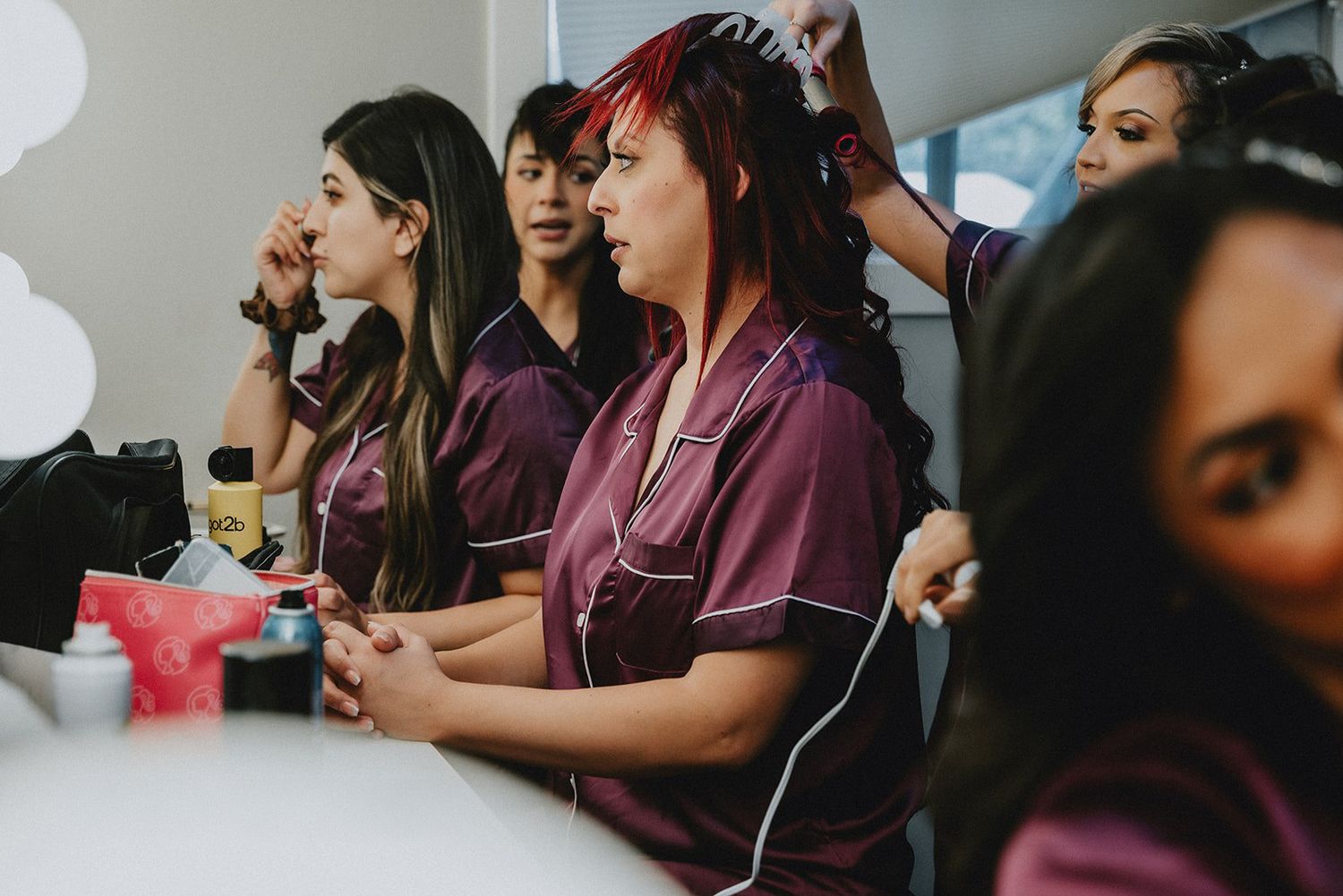 Bridesmaids in matching burgundy satin robes getting hair and makeup done together before a wedding ceremony.