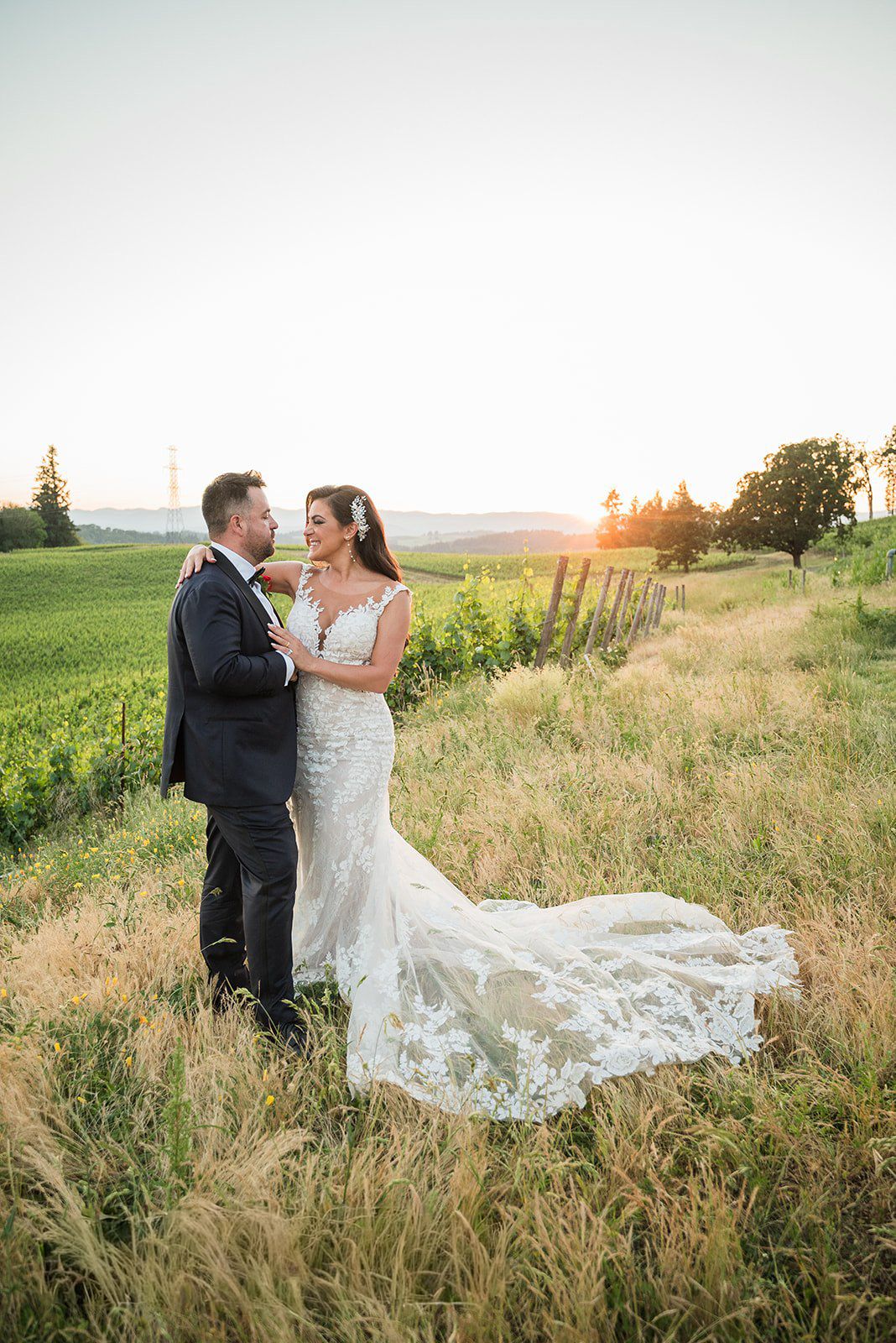 Bride and groom embrace in golden sunset vineyard, her lace mermaid gown trailing through tall grass.