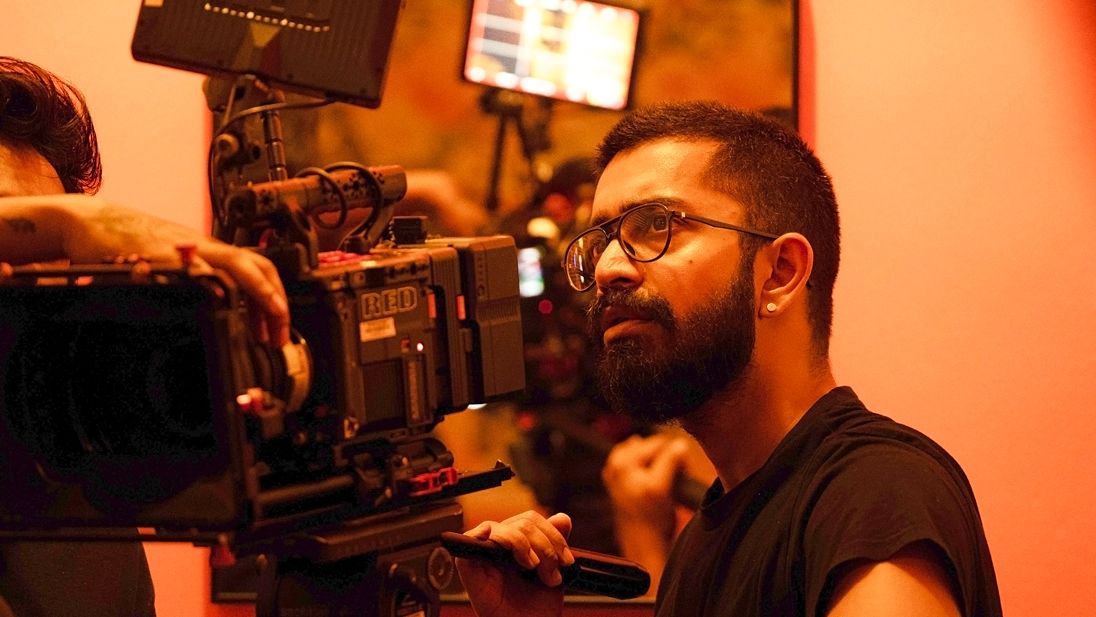 A bearded filmmaker operates a professional cinema camera on a film set bathed in warm orange lighting.