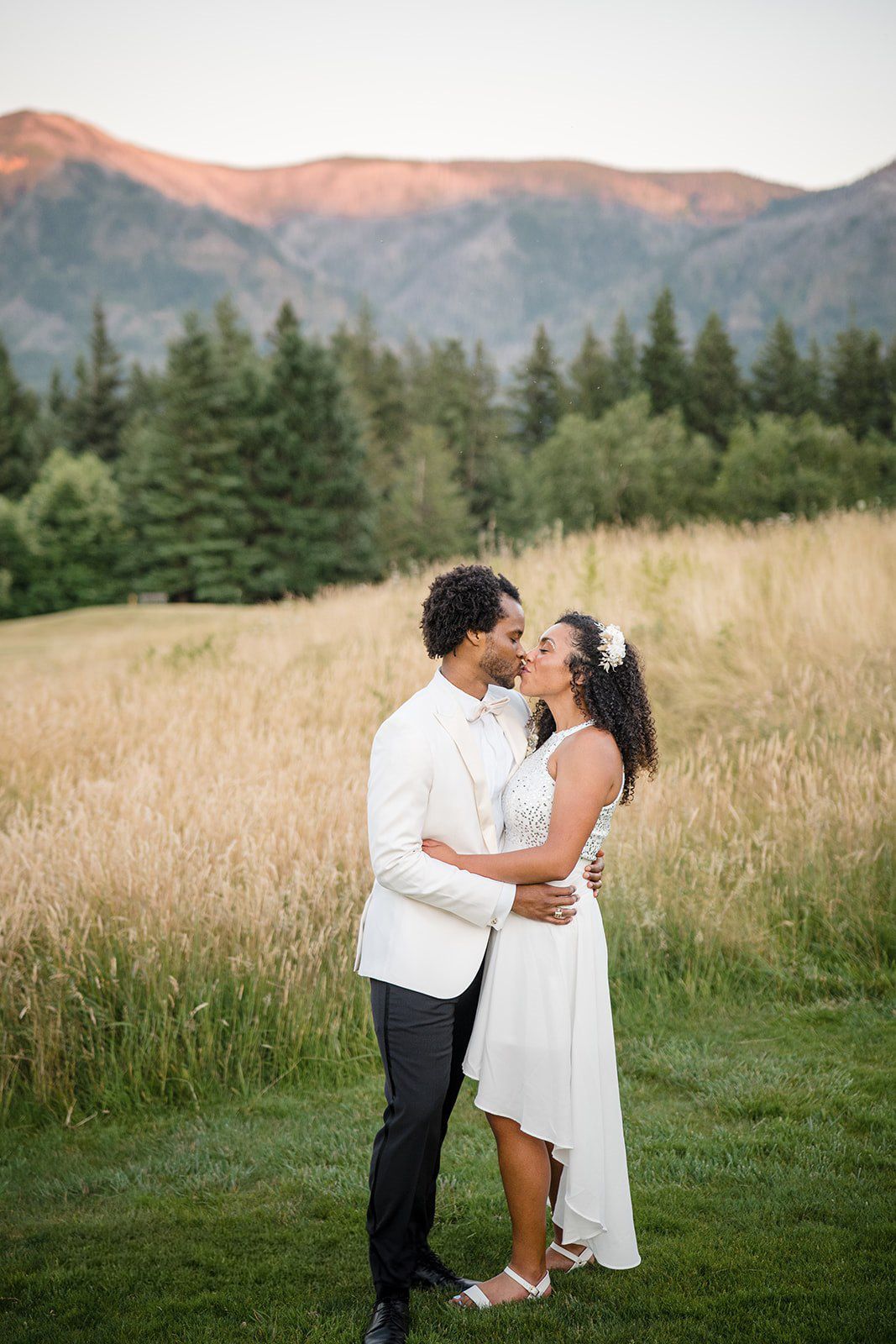 Bride and groom share a kiss at sunset with mountain backdrop and golden meadow at outdoor wedding.