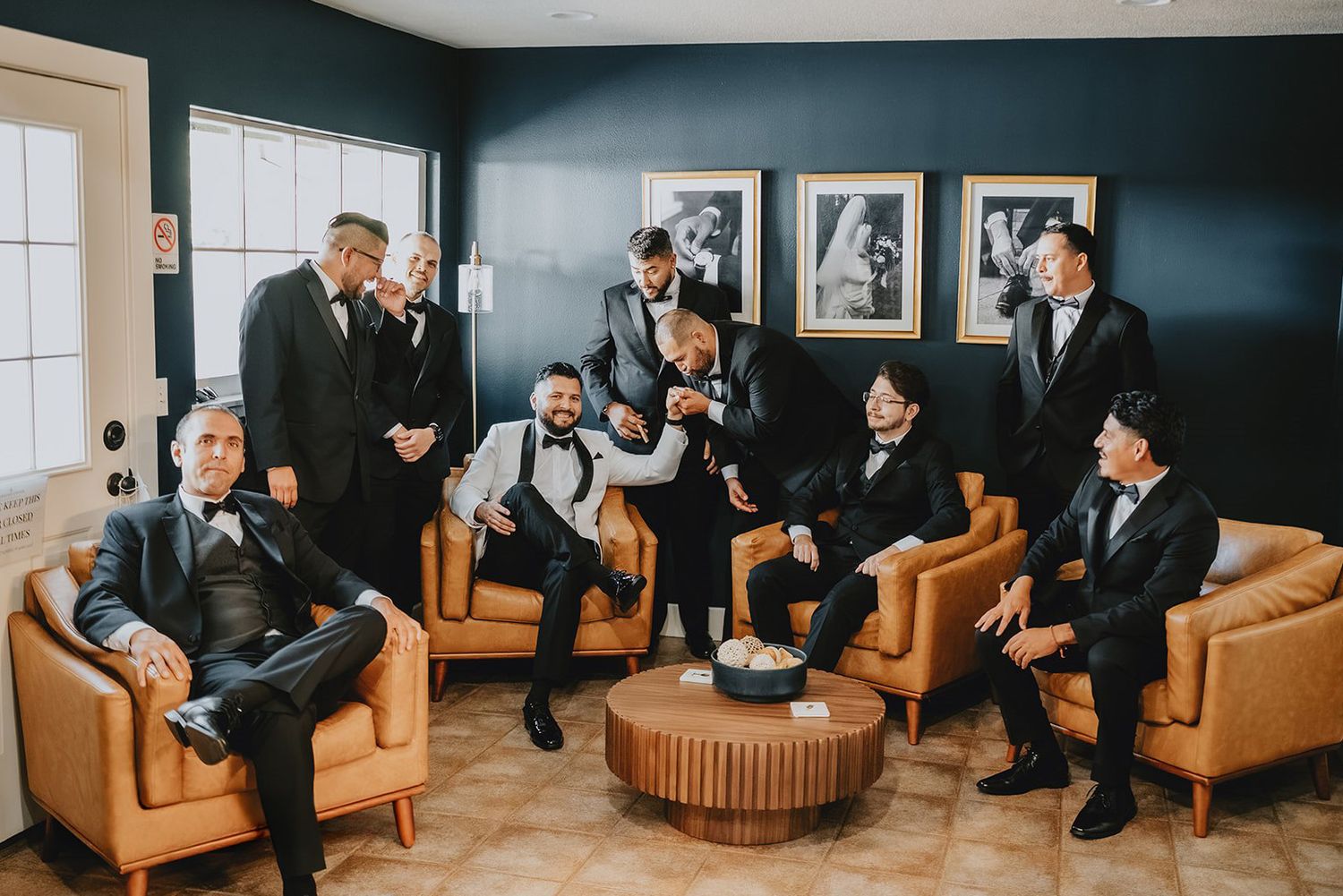 A groom and his groomsmen in black tuxedos gather and laugh together in a stylish lounge room before the wedding.