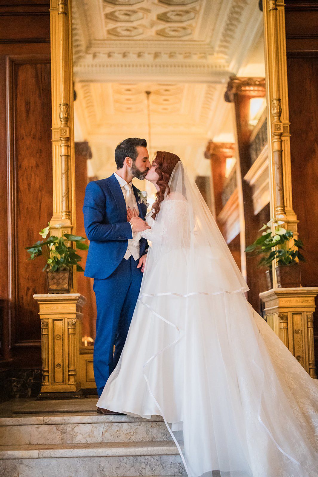 Newlyweds share a kiss in grand ornate hall, bride in flowing tiered veil, groom in blue suit.