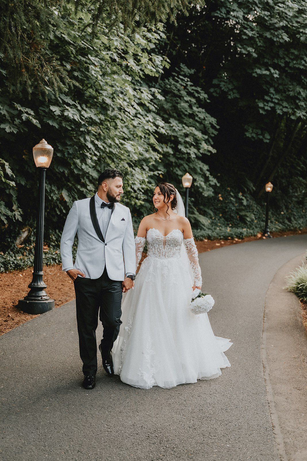Bride and groom walk hand-in-hand down a tree-lined path, she in a lace ballgown, he in a white tuxedo jacket.