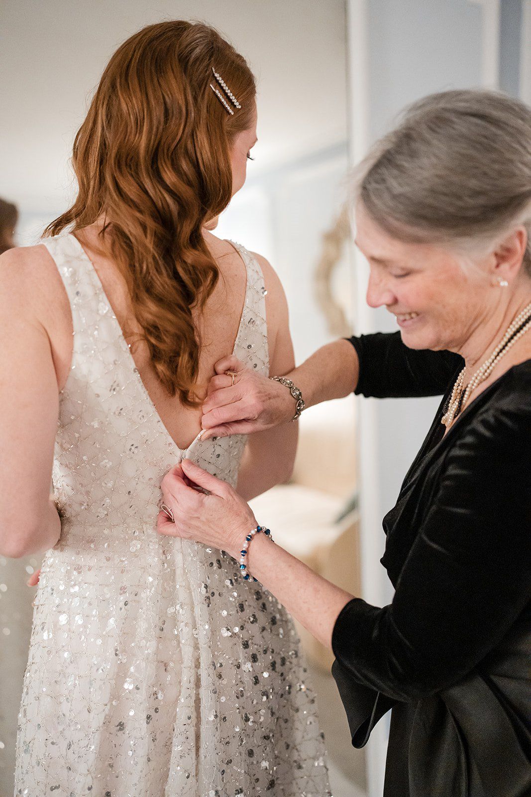 Older woman helping bride button up sparkly wedding dress from behind, smiling warmly in bright room.