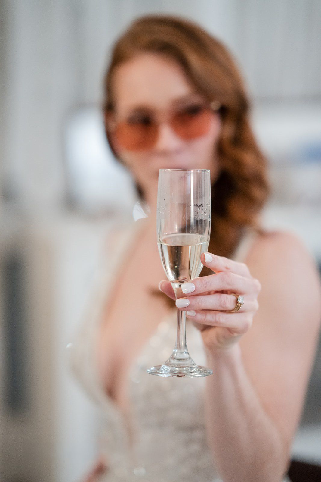 Bride in sunglasses holding a champagne flute toward camera, wearing a white dress and ring.