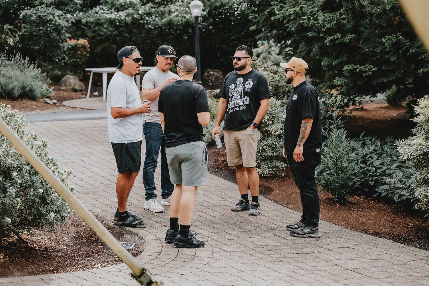 Five men in casual attire conversing outdoors on a stone pathway surrounded by lush green garden landscaping.