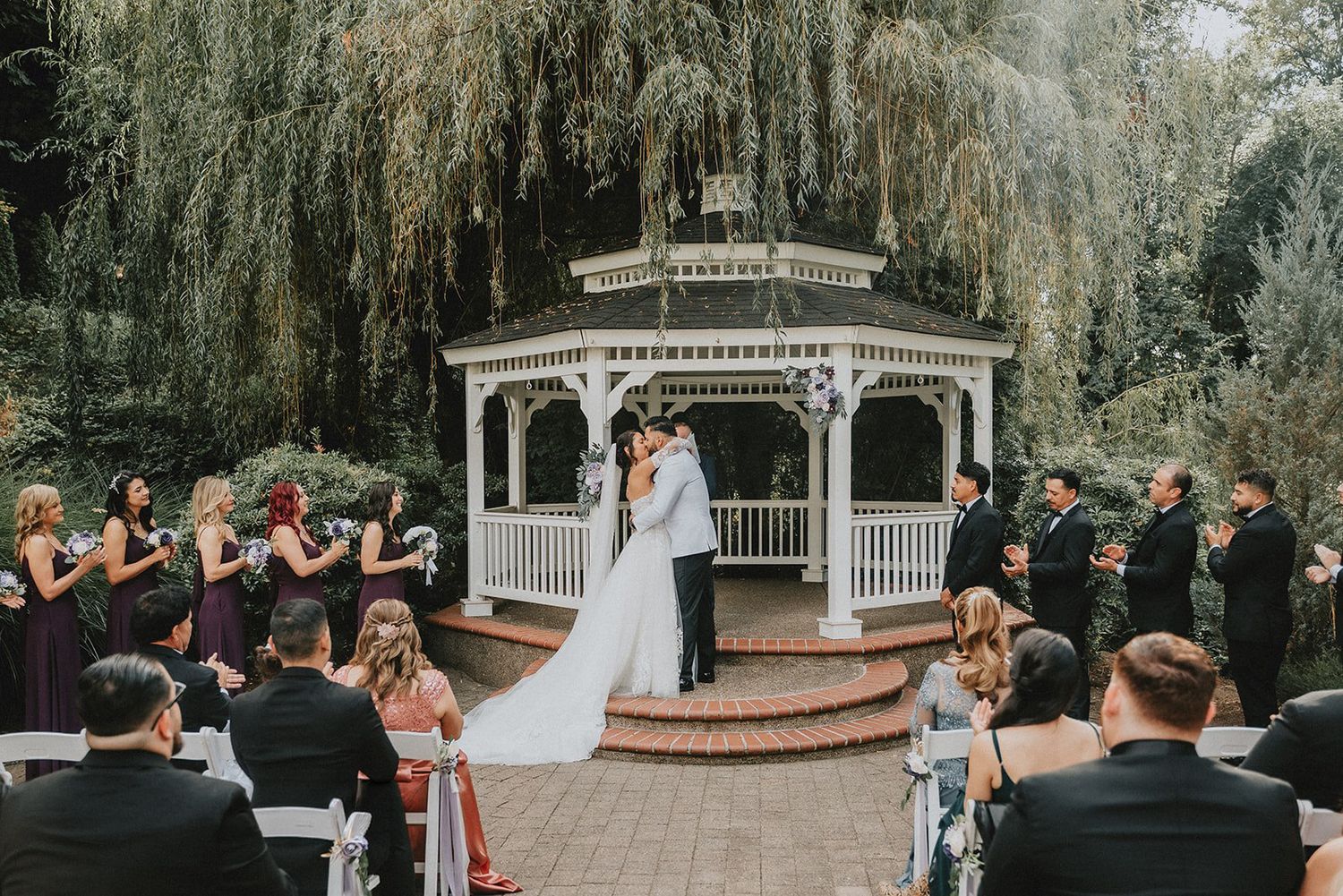 Bride and groom share first kiss at outdoor gazebo wedding ceremony surrounded by guests and lush weeping willow trees.