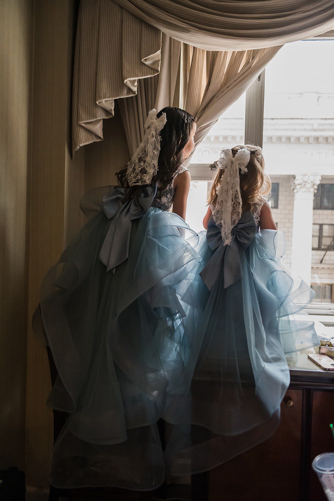 Two flower girls in blue ruffle dresses with large bows and lace headpieces gaze out a window.