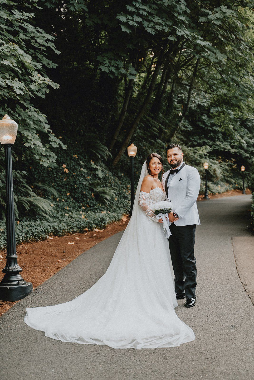 Bride in white gown with long train and groom in white tuxedo jacket pose on a tree-lined path.