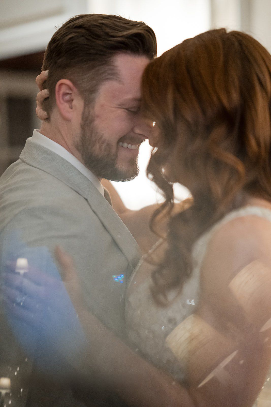 Smiling groom in gray suit shares intimate forehead touch with bride during romantic wedding first dance.