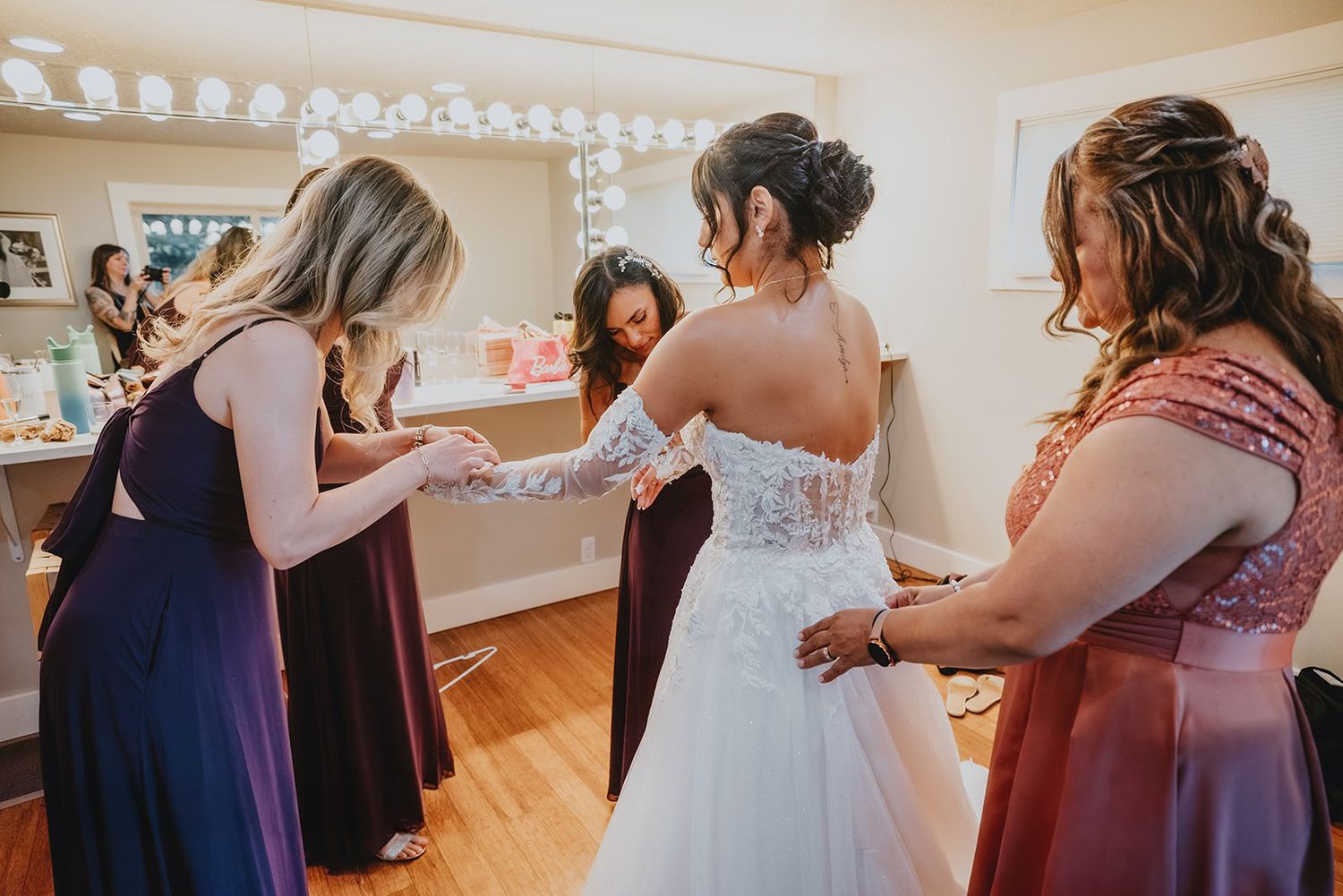 Bridesmaids helping bride into white lace wedding gown in bridal suite with Hollywood mirror.