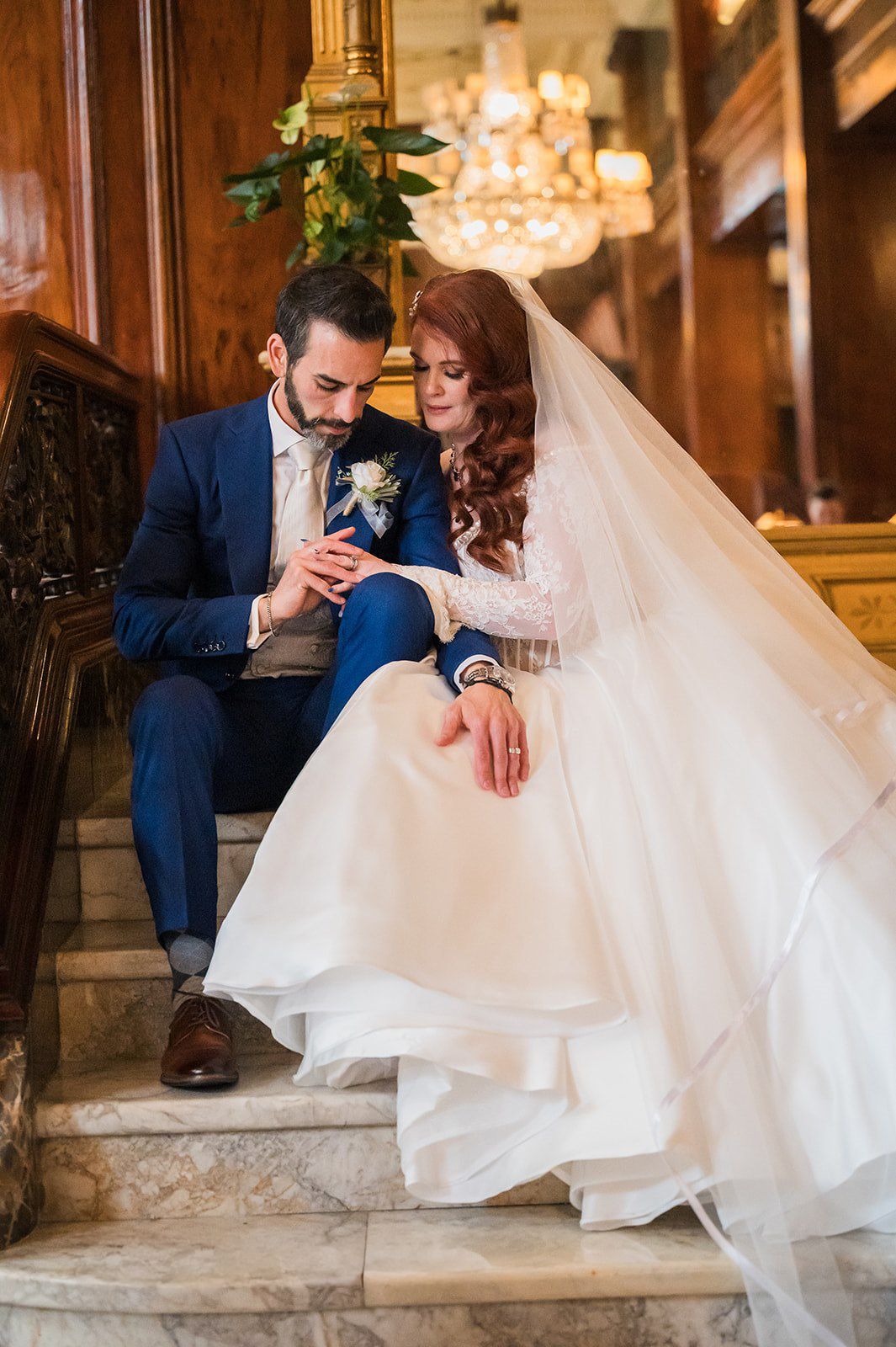 Bride and groom in blue suit sit on marble stairs in elegant venue, gazing at wedding rings.