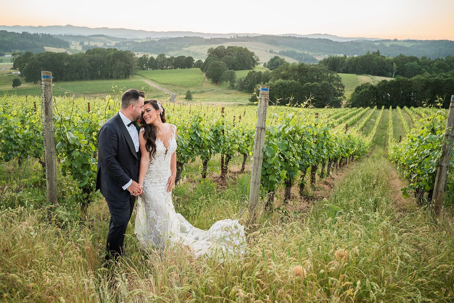 Bride and groom embrace romantically in a lush vineyard at sunset, surrounded by rolling green hills.