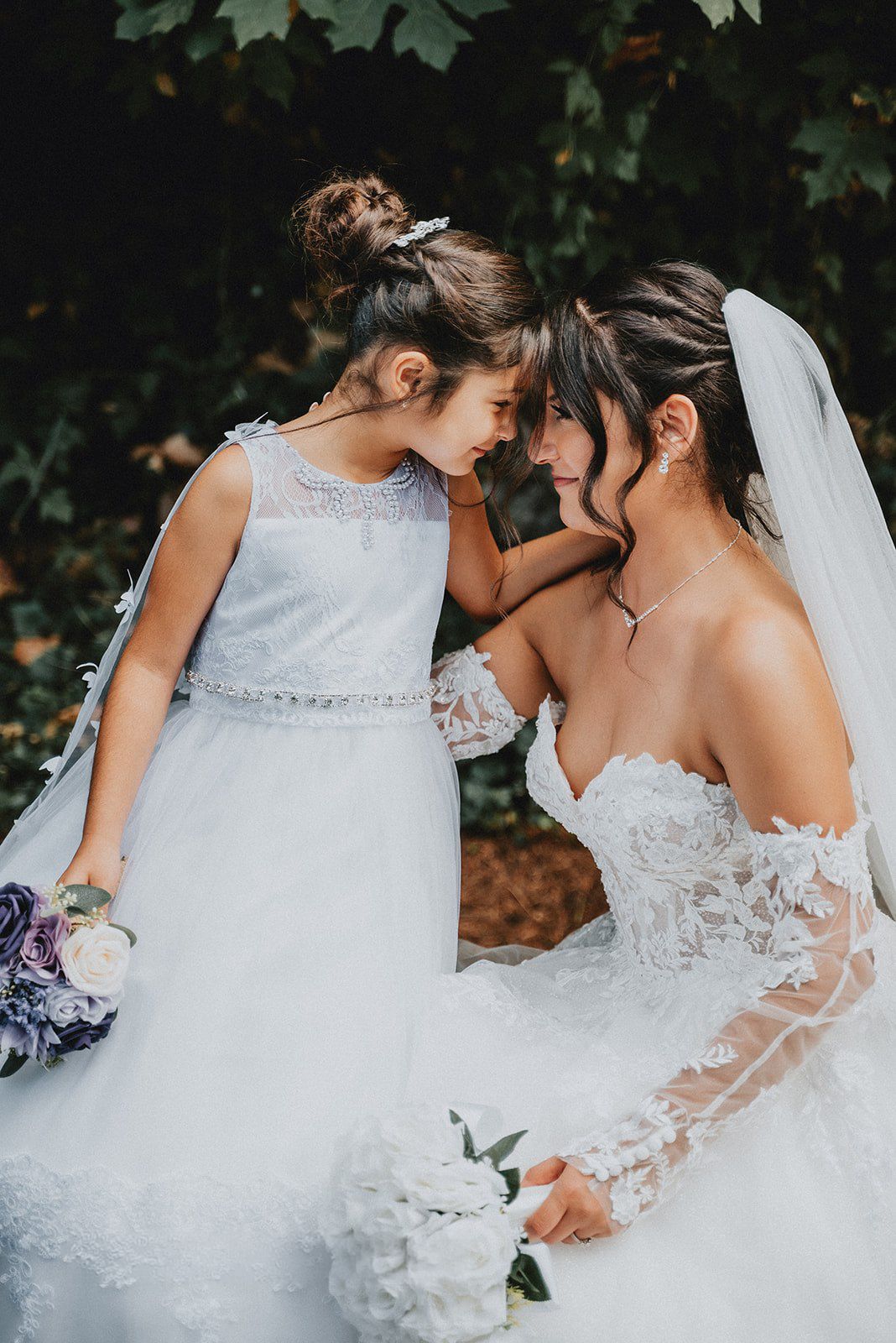 Bride in lace wedding gown shares tender forehead touch with flower girl holding white bouquet outdoors.