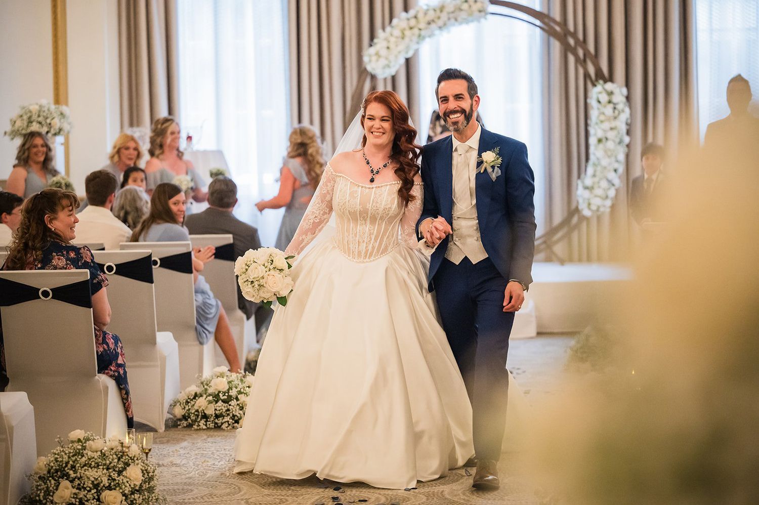 Bride and groom walking down the aisle at an elegant indoor wedding ceremony with floral arch and white decorations.
