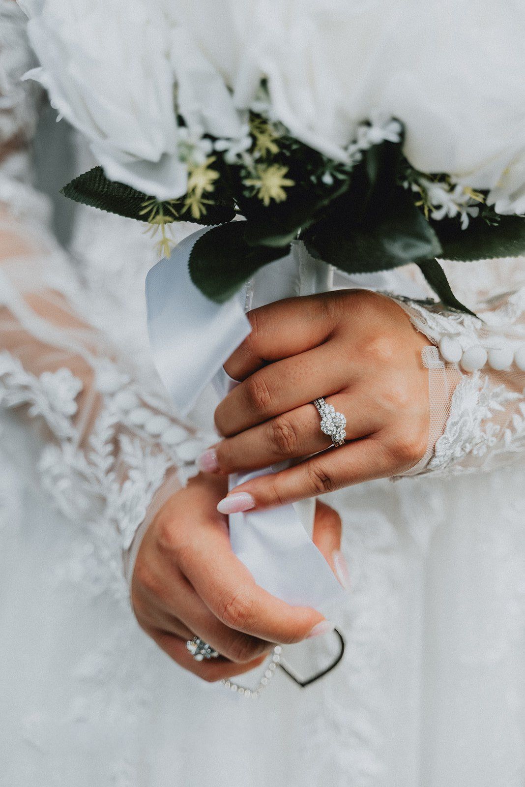 Bride holding white floral bouquet with ribbon, showcasing diamond engagement ring on lace wedding dress sleeve.