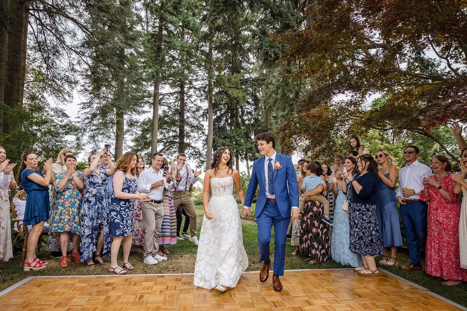Bride and groom walk hand-in-hand on outdoor dance floor surrounded by cheering guests at a garden wedding.
