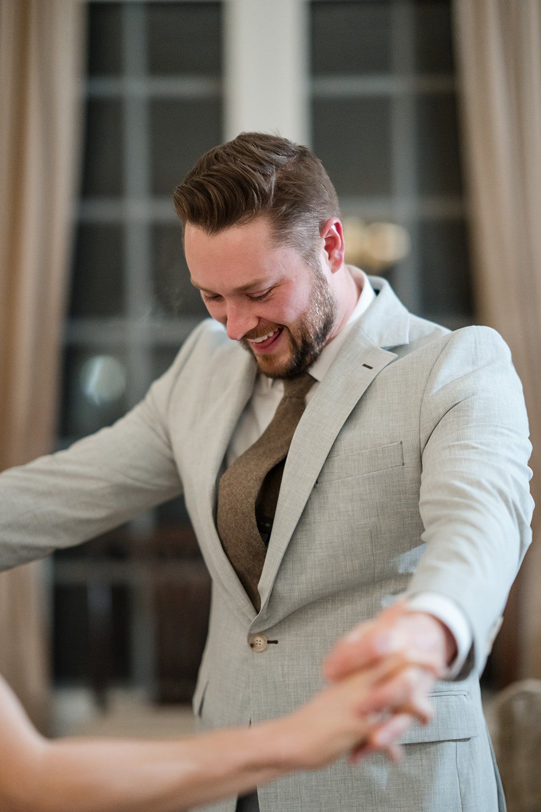 Smiling groom in gray suit holds hands with bride, dancing joyfully at elegant indoor wedding venue.