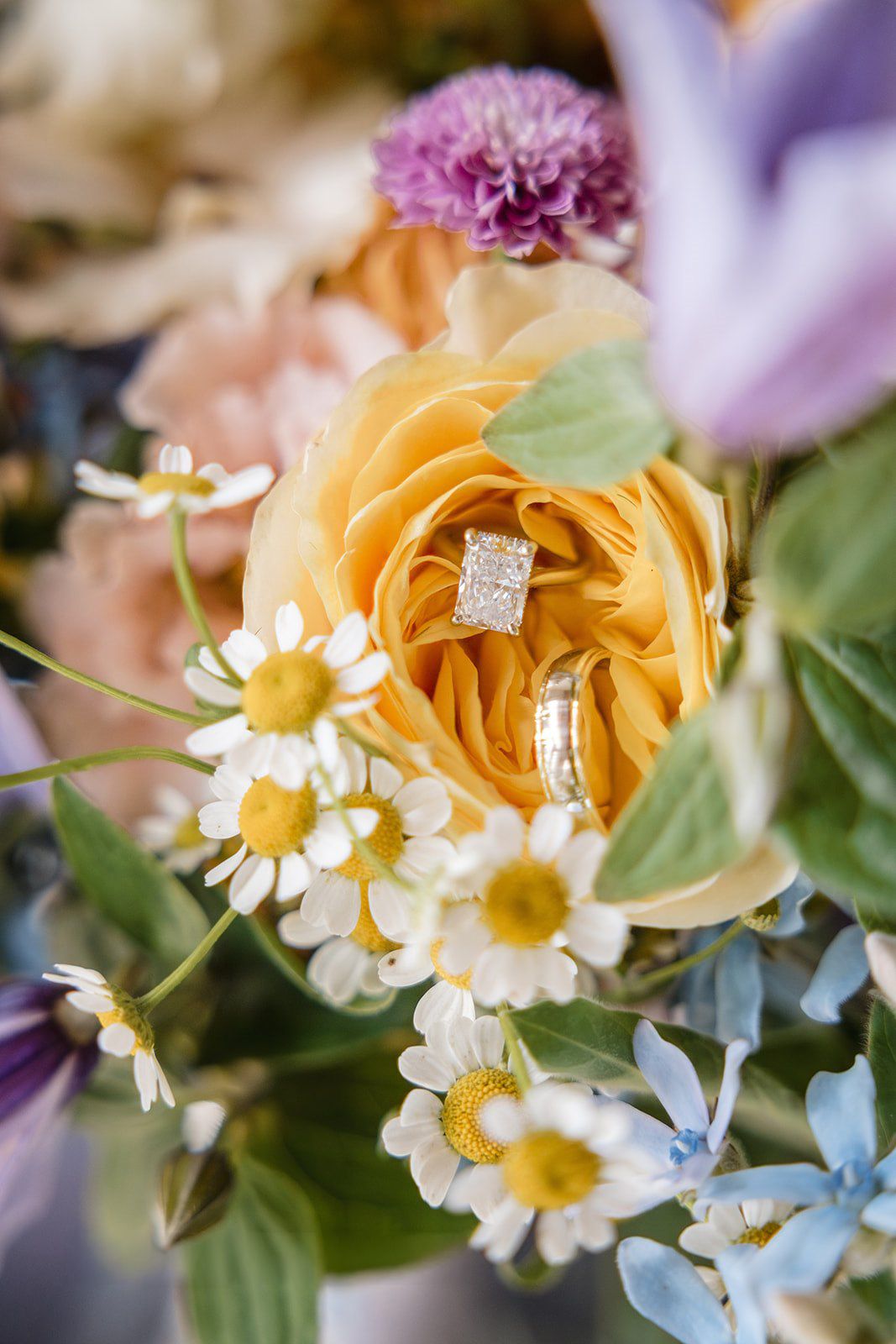Diamond engagement ring and gold wedding band nestled in vibrant yellow rose surrounded by colorful wildflower bouquet.
