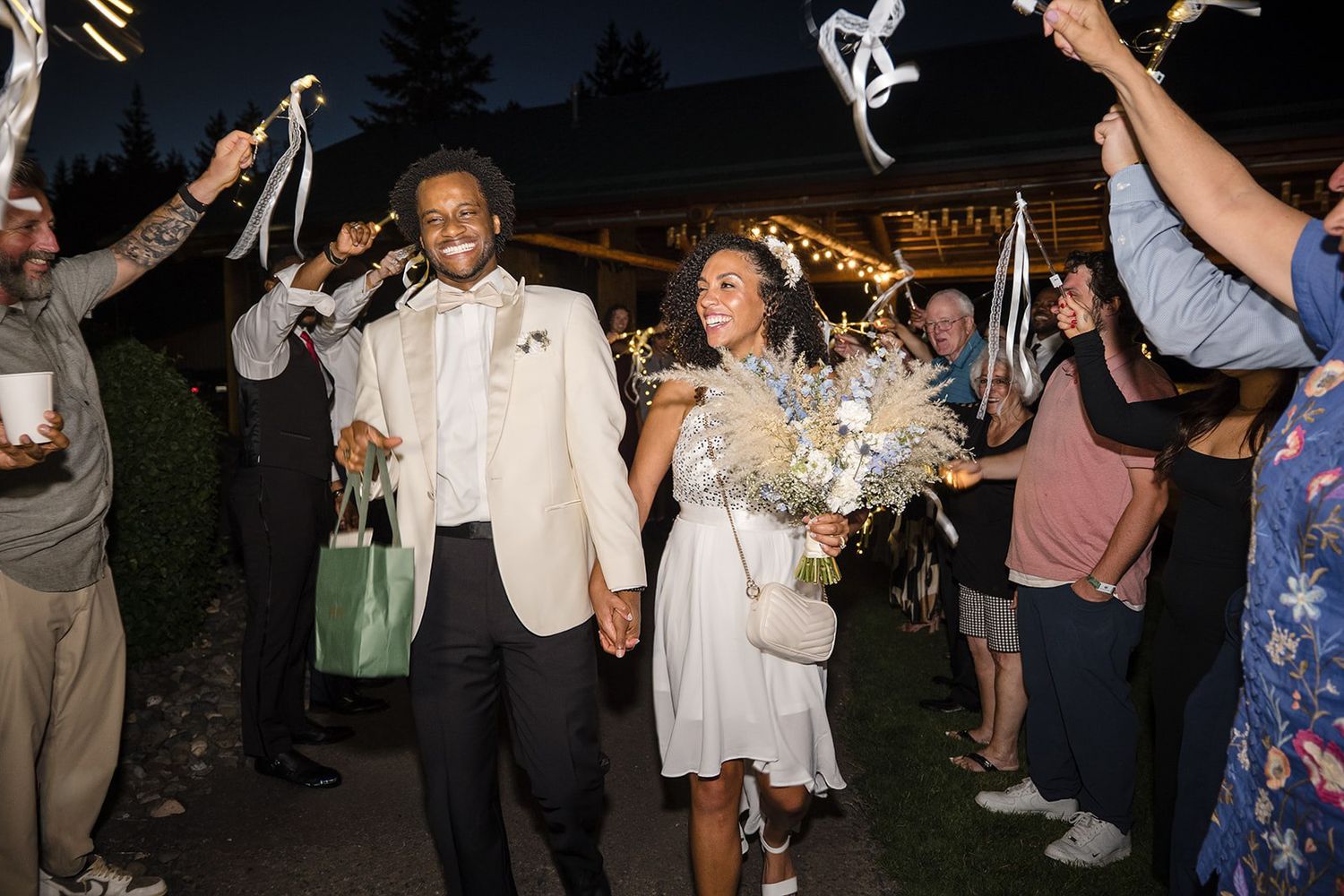 Newlywed couple walks through sparkler exit at outdoor wedding reception at night, bride holding bouquet.