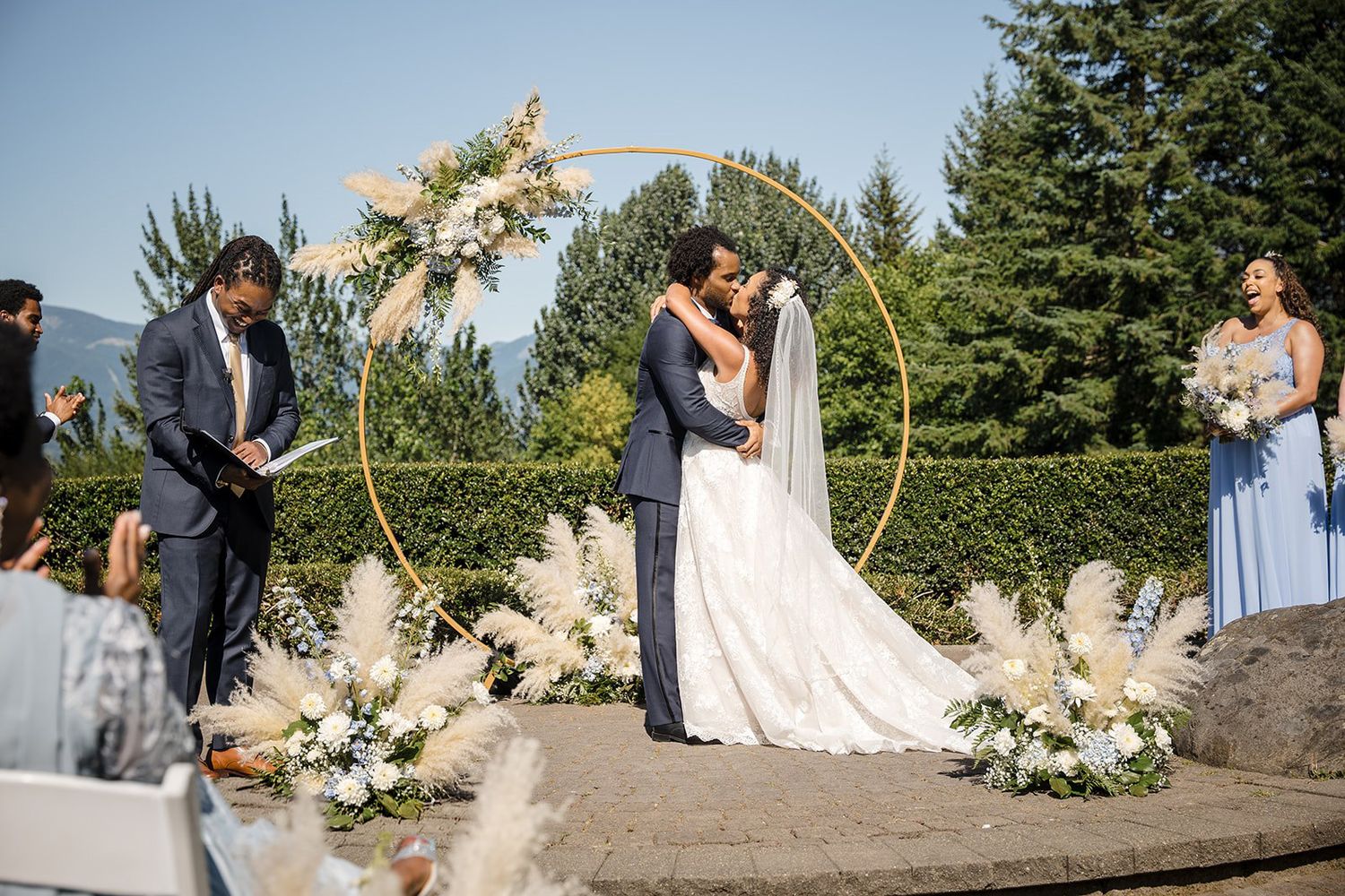 Bride and groom share first kiss at outdoor wedding ceremony under golden hoop arch with pampas grass floral arrangements.