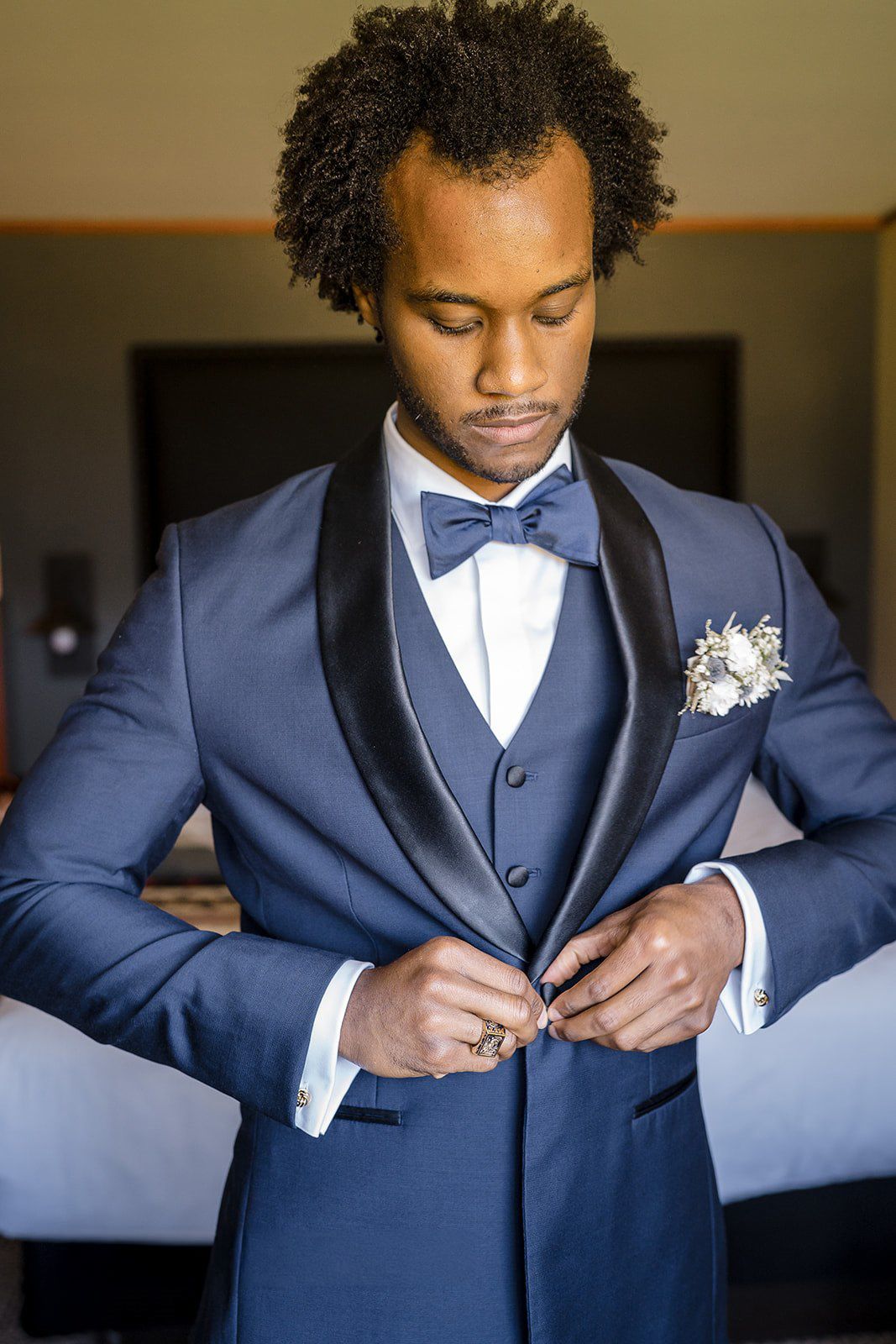 A groom in a blue tuxedo with black lapels and bow tie buttons his jacket while wearing a floral boutonniere.