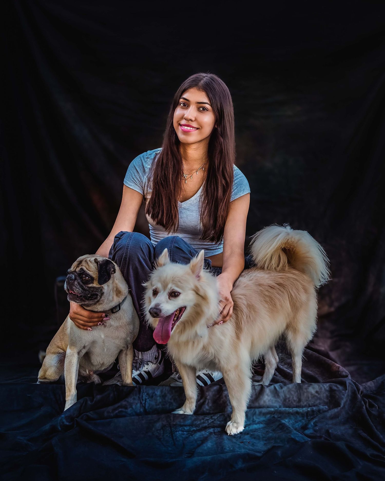 A young teenager with straight hair sitting down with a Pug and another white mixed breed dog against a dark textured backdrop.