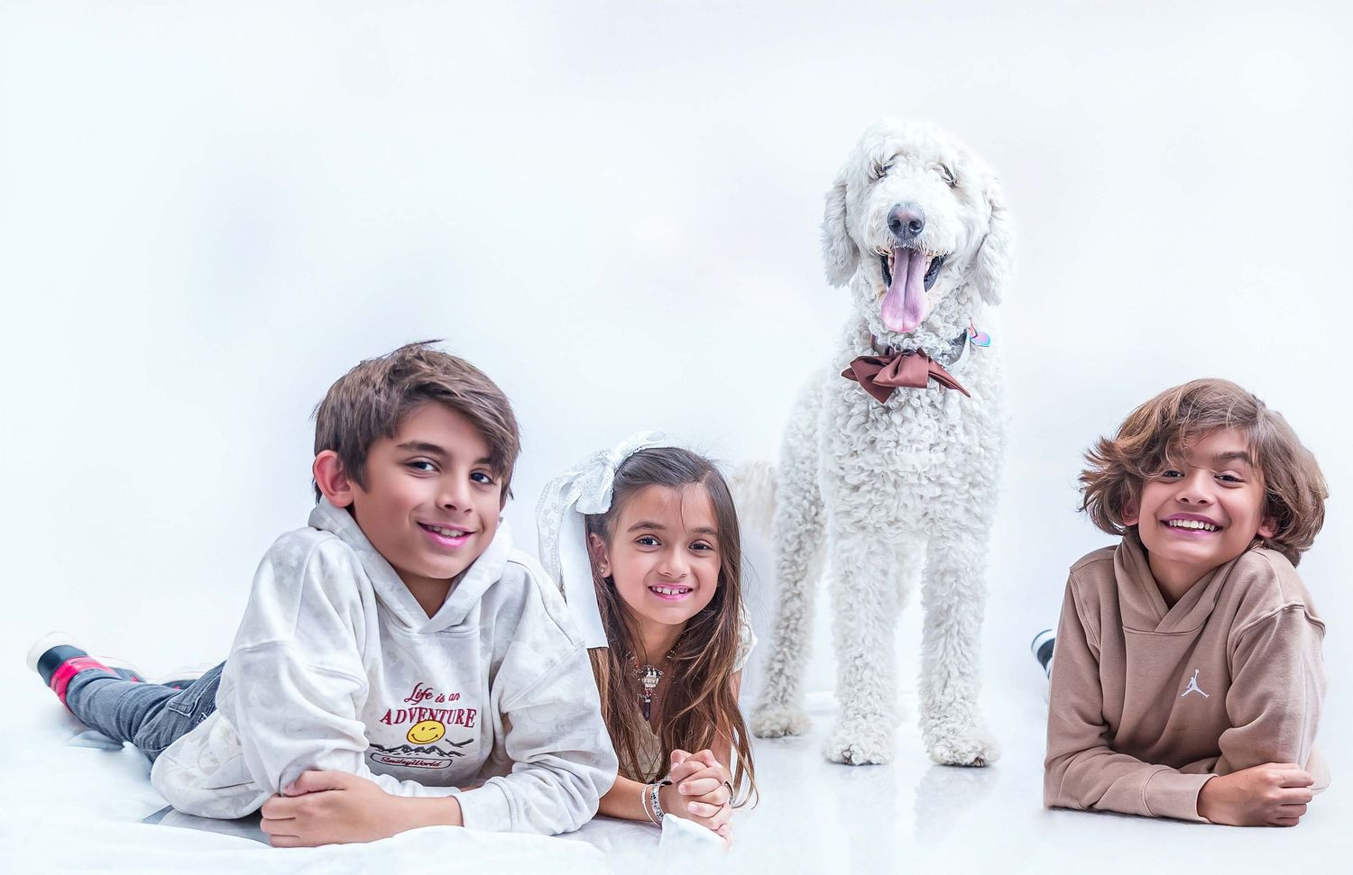 Three smiling children posing lying down with a fluffy white Standard Poodle wearing a bow tie in a professional studio photography session.