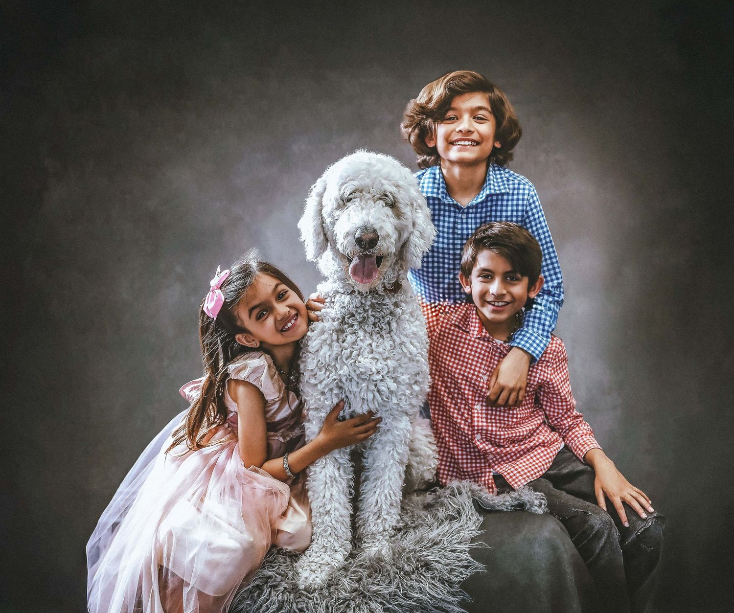 Three smiling children hugging a large, fluffy white Standard Poodle during a professional studio photography session.