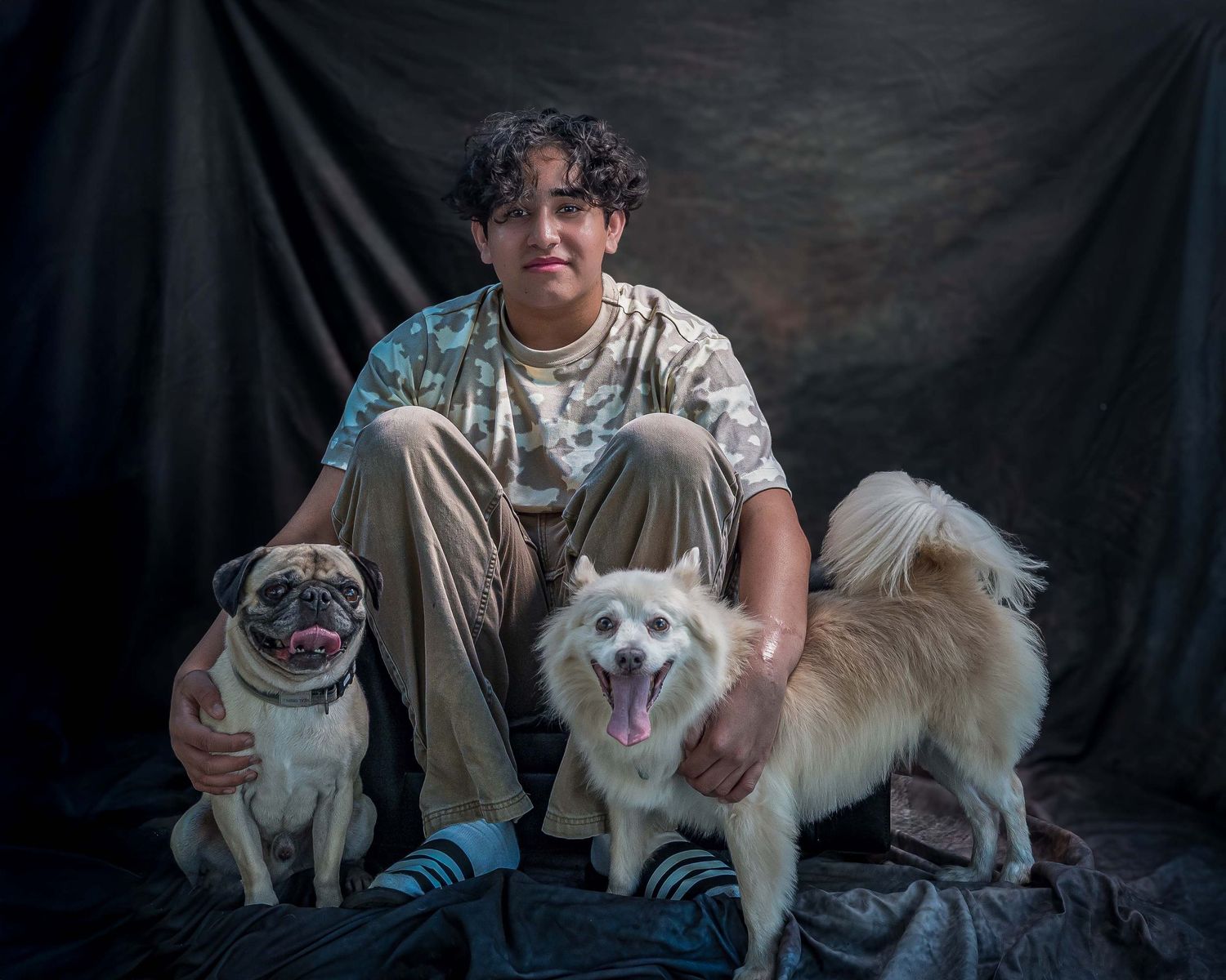 A young boy with curly hair sitting down with a Pug and another white mixed breed dog against a dark textured backdrop.