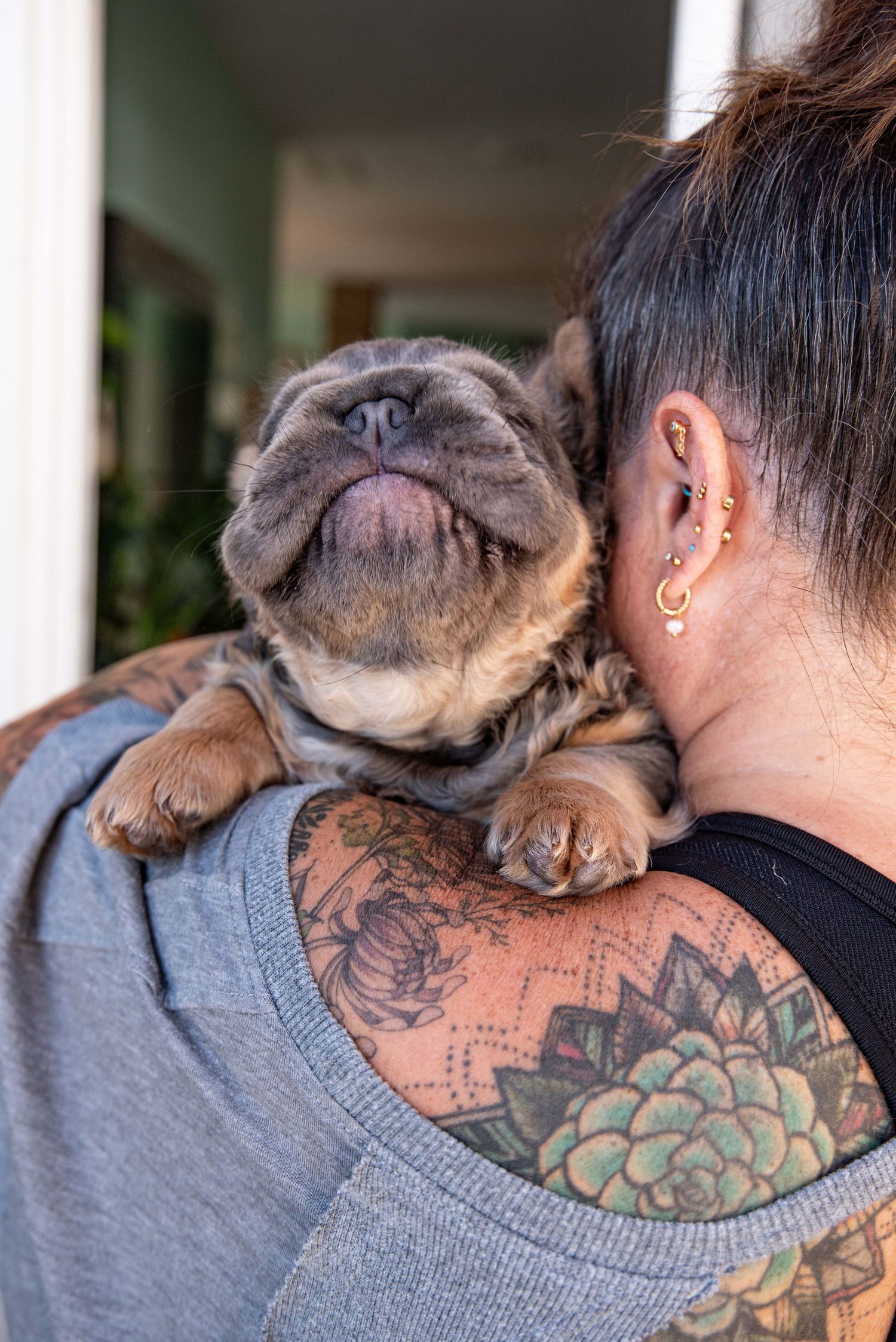 Close-up of a French bulldog puppy cuddling with a woman featuring floral shoulder tattoos and gold ear piercings.