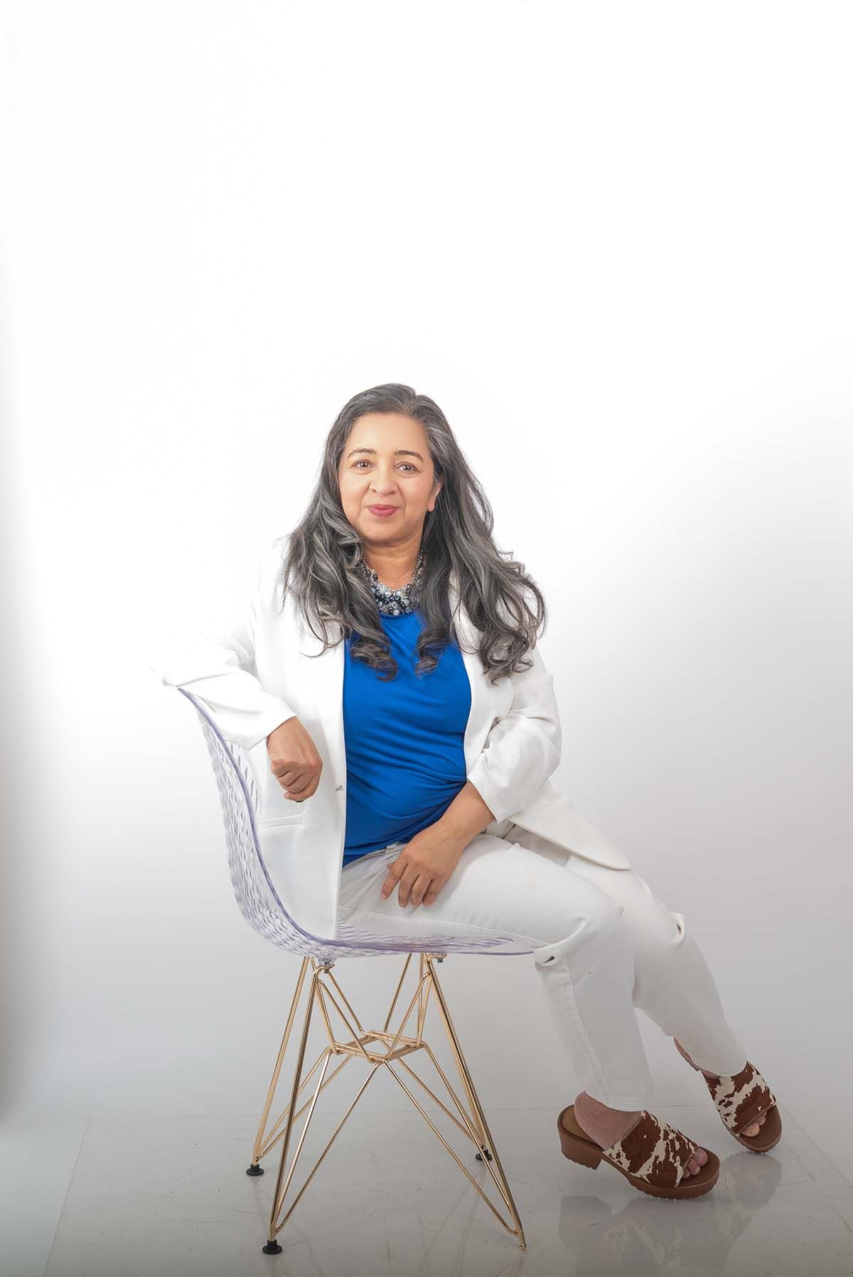 High-key studio portrait of Anu Kalle wearing a white blazer and blue top, sitting in a clear modern chair on a white background.