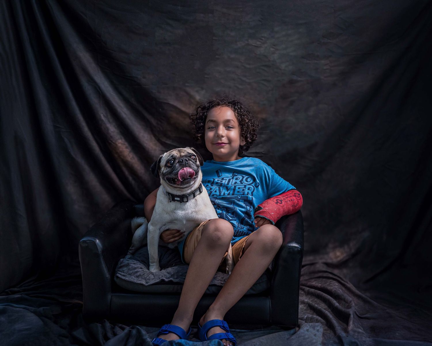 A young boy with curly hair sitting in a small black armchair with a Pug dog against a dark textured backdrop.