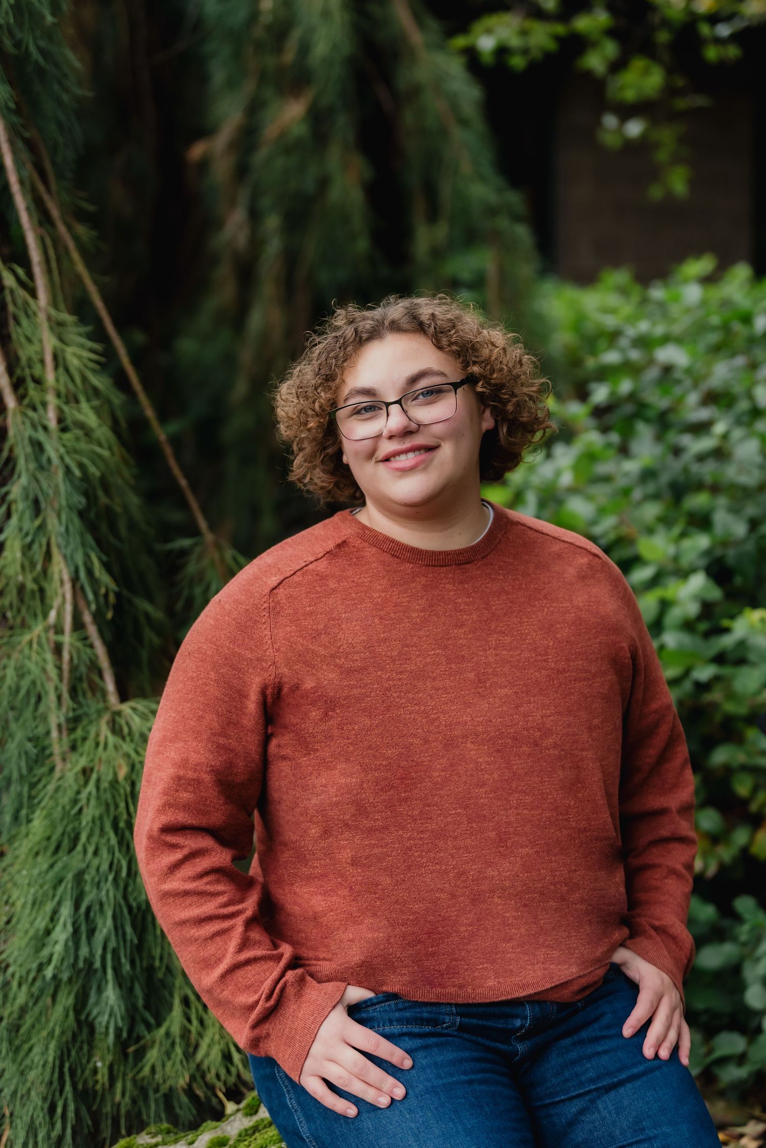 A smiling figure in glasses wearing a rust-colored sweater and jeans poses against a green outdoor backdrop.