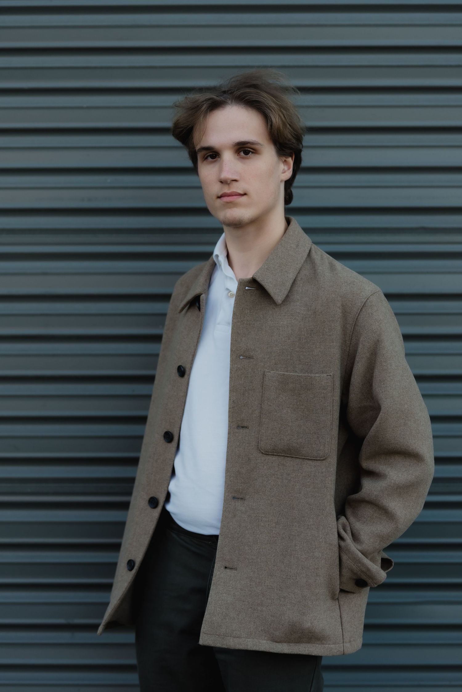 A figure poses in a brown jacket and white shirt against a corrugated metal wall background.