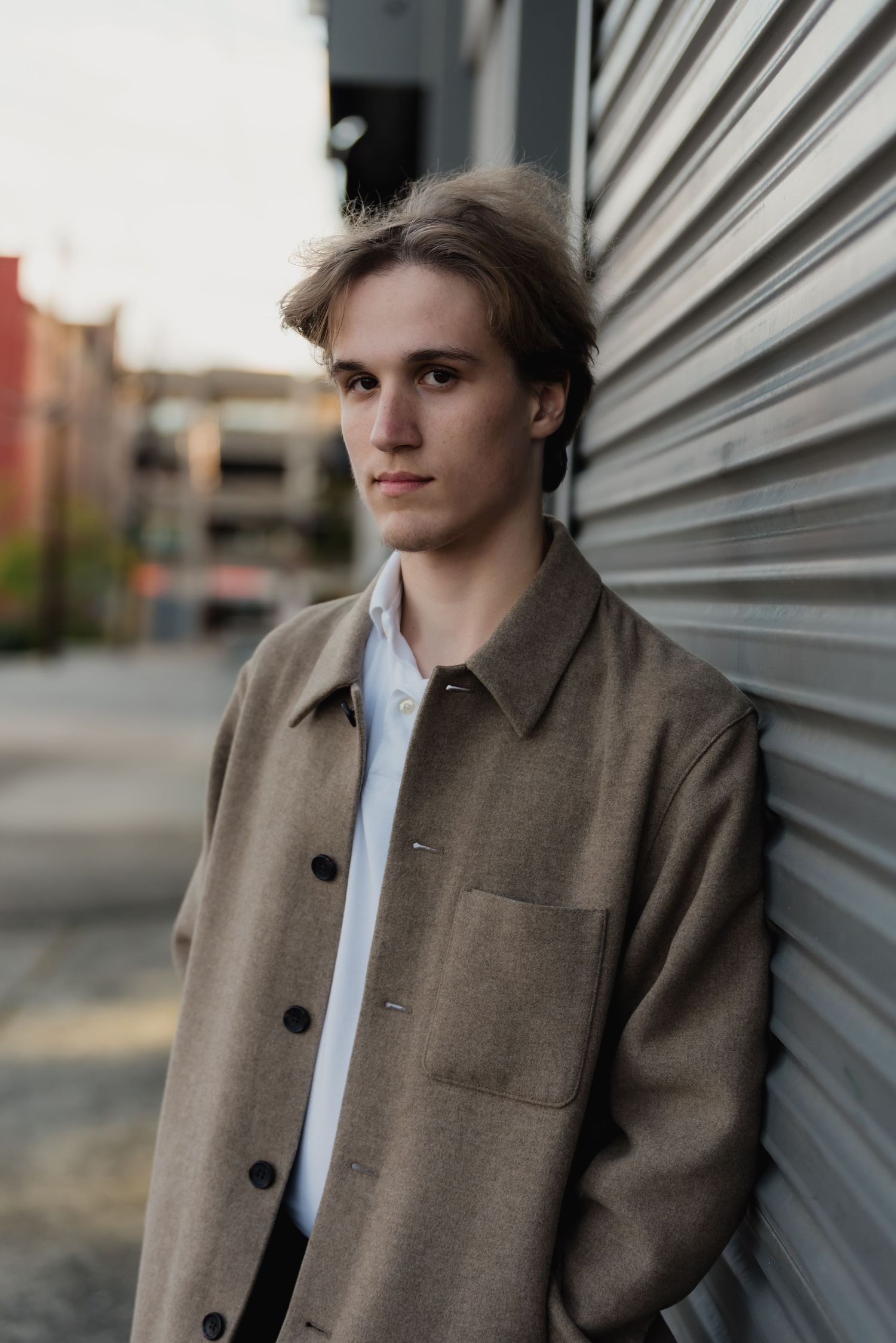 A high school senior boy in a brown coat leans against corrugated metal wall in moody urban outdoor portrait in Everett, Washington.