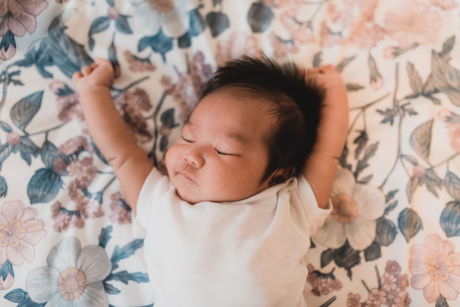 Sleeping baby lies on floral patterned bedding in soft pink and blue tones.