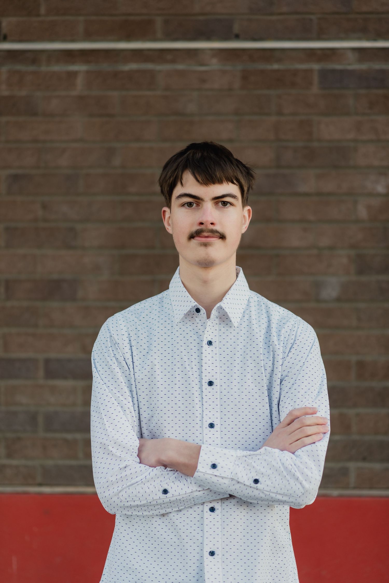 Person in white polka dot button-down shirt with mustache stands with arms crossed against brick wall with red accent.