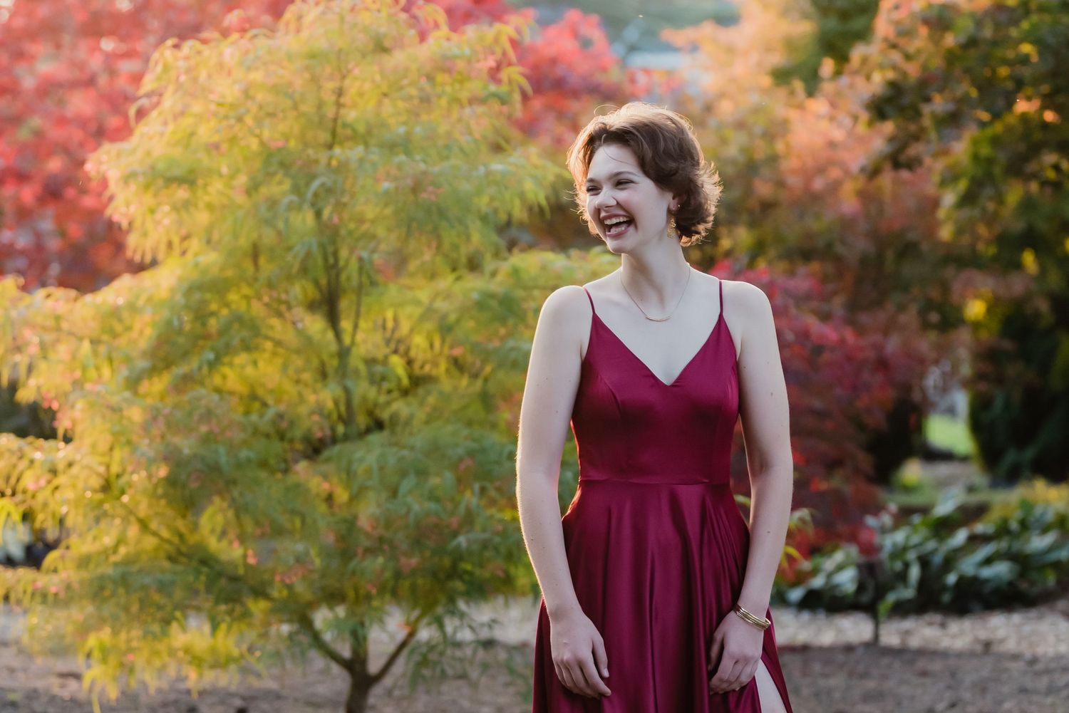 Woman in burgundy dress poses against vibrant autumn foliage with yellow and orange trees in background.